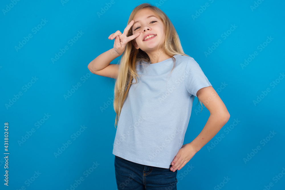 Fototapeta premium beautiful Caucasian little girl wearing blue T-shirt over blue background making v-sign near eyes. Leisure, coquettish, celebration, and flirt concept.