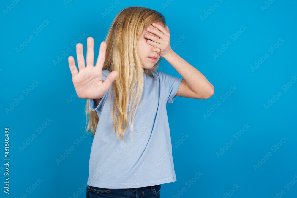 beautiful Caucasian little girl wearing blue T-shirt over blue background covers eyes with palm and doing stop gesture, tries to hide. Don't look at me, I don't want to see, feels ashamed or scared. beautiful Caucasian little girl wearing blue T-shirt over blue background covers eyes with palm and doing stop gesture, tries to hide. Don't look at me, I don't want to see, feels ashamed or scared.