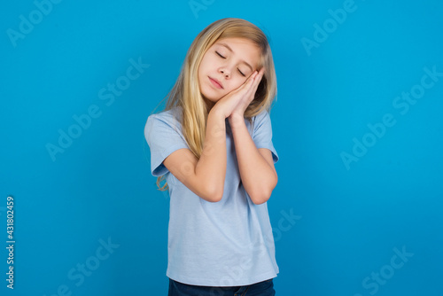 Relax and sleep time. Tired beautiful Caucasian little girl wearing blue T-shirt over blue background with closed eyes leaning on palms making sleeping gesture.