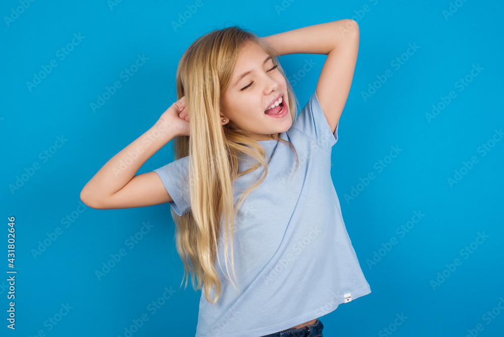 beautiful Caucasian little girl wearing blue T-shirt over blue background relaxing and stretching, arms and hands behind head and neck smiling happy