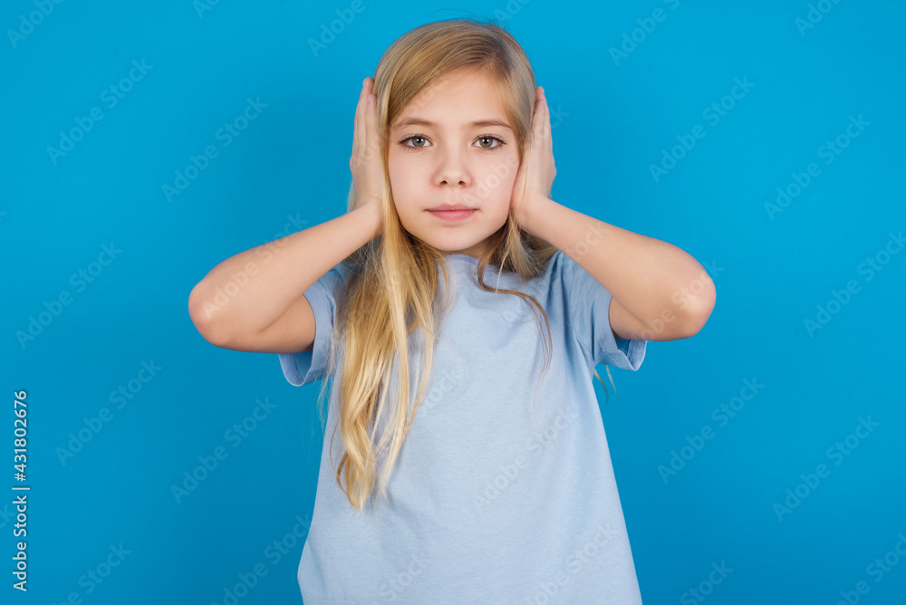 Fototapeta premium Frustrated beautiful Caucasian little girl wearing blue T-shirt over blue background plugging ears with hands does not wanting to listen hard rock, noise or loud music.