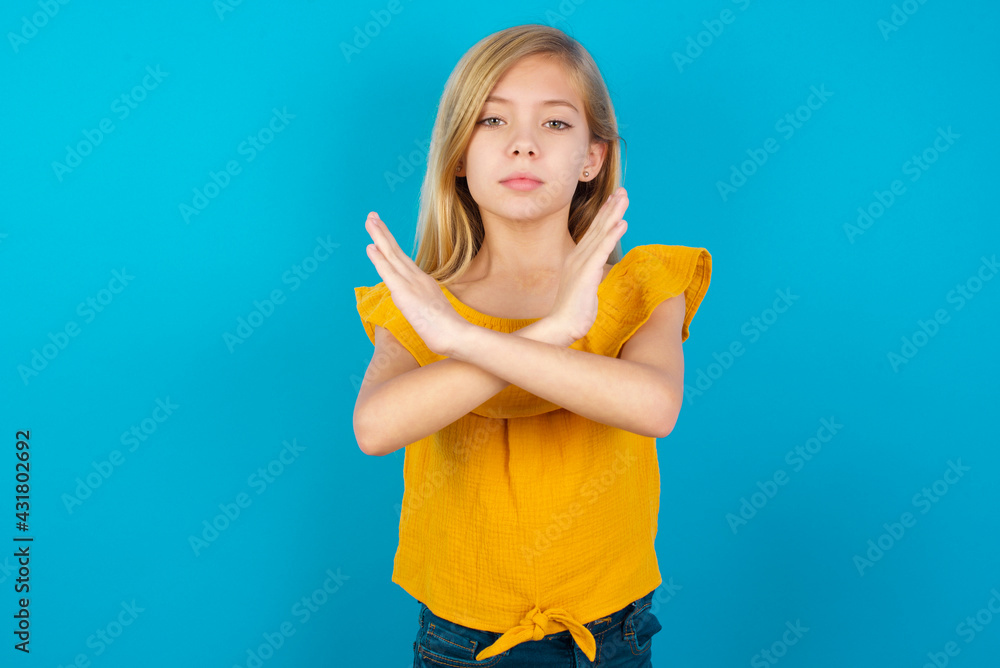 Caucasian kid girl wearing yellow T-shirt against blue wall Rejection expression crossing arms doing negative sign, angry face Caucasian kid girl wearing yellow T-shirt against blue wall Rejection expression crossing arms doing negative sign, angry face