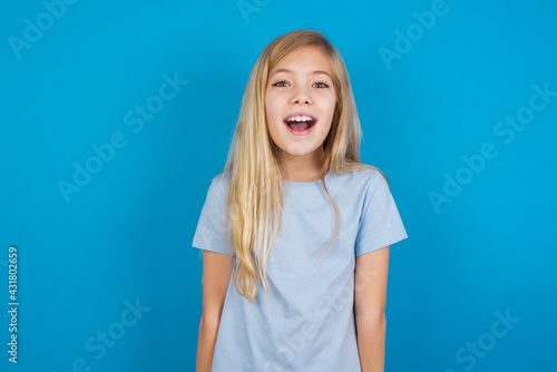 Surprised beautiful Caucasian little girl wearing blue T-shirt over blue background, shrugs shoulders, looking sideways, being happy and excited. Sudden reactions concept. © Jihan