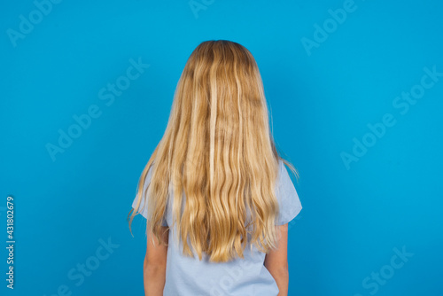 beautiful Caucasian little girl wearing blue T-shirt over blue background standing backwards looking away with arms on body.