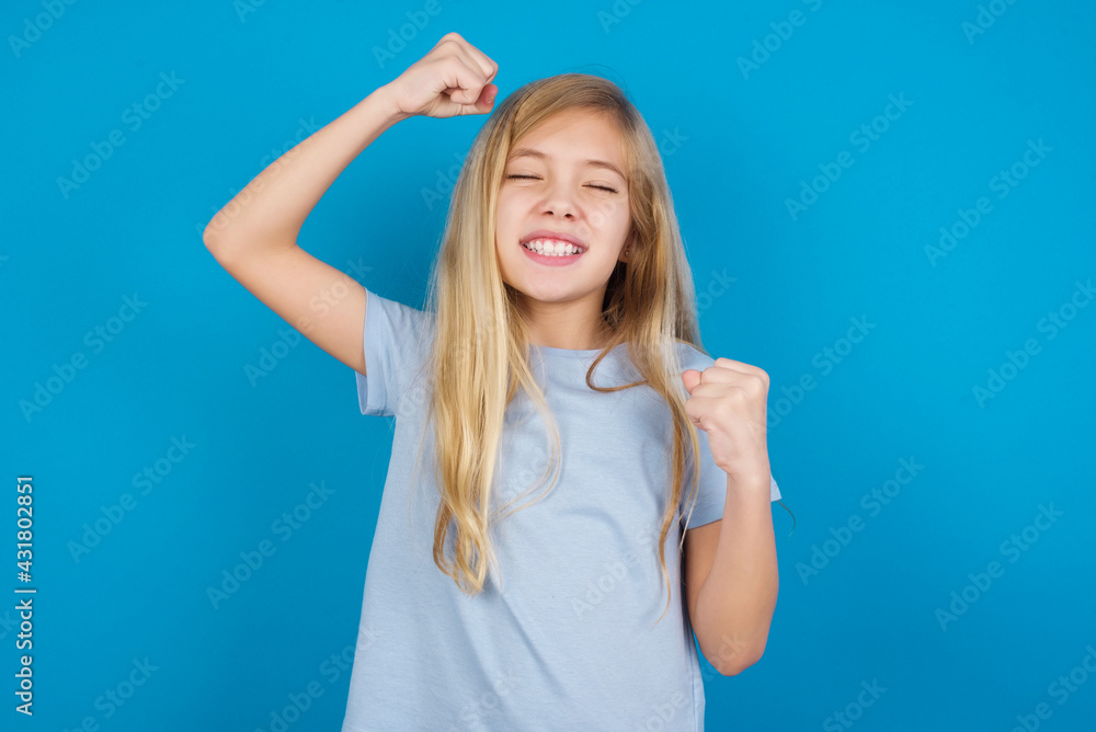Fototapeta premium Attractive beautiful Caucasian little girl wearing blue T-shirt over blue background celebrating a victory punching the air with his fists and a beaming toothy smile.