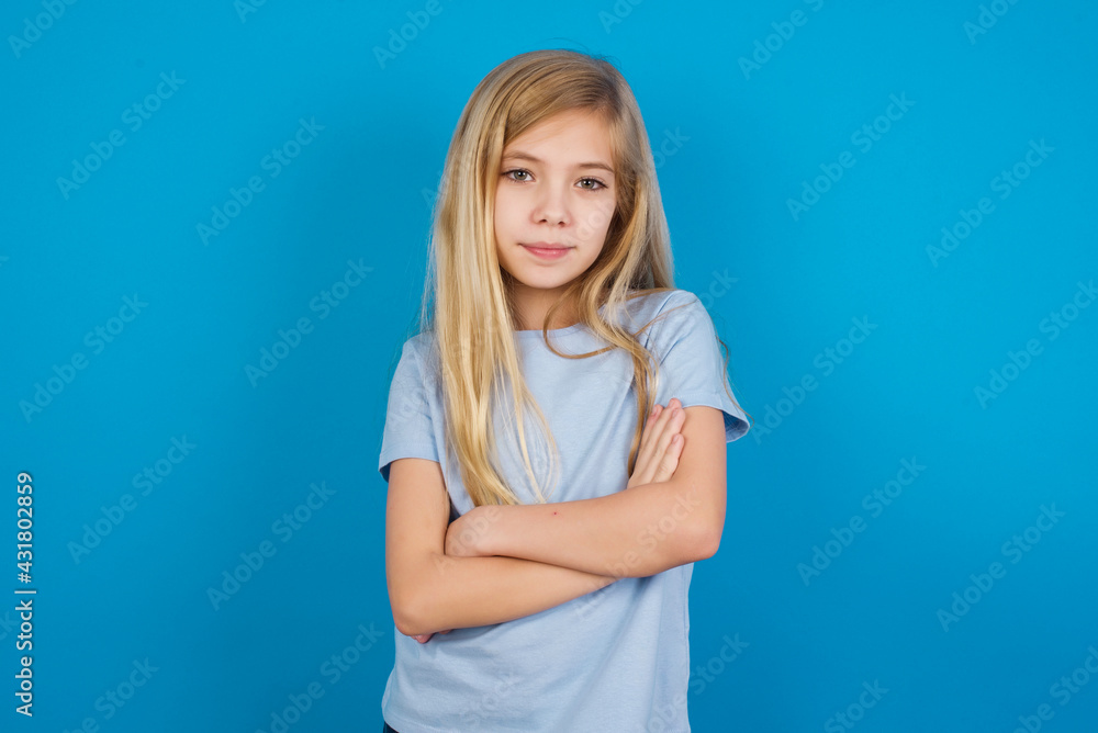 Waist up shot of self confident beautiful Caucasian little girl wearing blue T-shirt over blue background has broad smile, crosses arms, happy to meet with colleagues. Waist up shot of self confident beautiful Caucasian little girl wearing blue T-shirt over blue background has broad smile, crosses arms, happy to meet with colleagues.