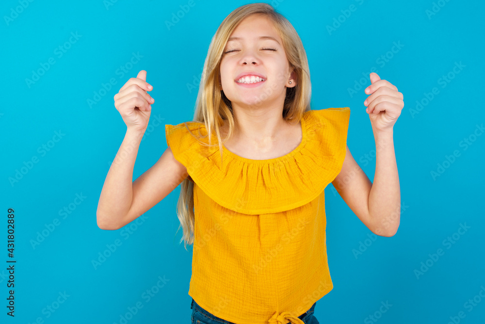 Caucasian kid girl wearing yellow T-shirt against blue wall being excited for success with raised arms and closed eyes celebrating victory. Winner concept. Caucasian kid girl wearing yellow T-shirt against blue wall being excited for success with raised arms and closed eyes celebrating victory. Winner concept.