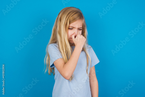 Foto beautiful Caucasian little girl wearing blue T-shirt over blue background, holding his nose because of a bad smell