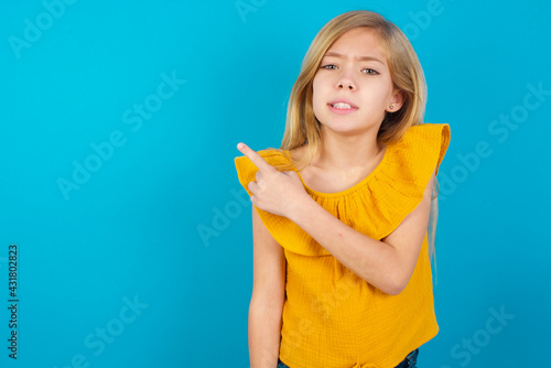 Caucasian kid girl wearing yellow T-shirt against blue wall Pointing aside worried and nervous with forefinger  concern and surprise concept.