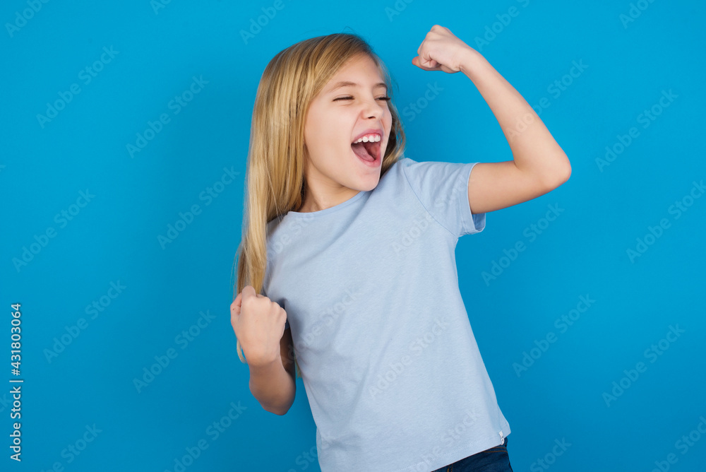 Fototapeta premium beautiful Caucasian little girl wearing blue T-shirt over blue background celebrating a victory punching the air with his fists and a beaming toothy smile
