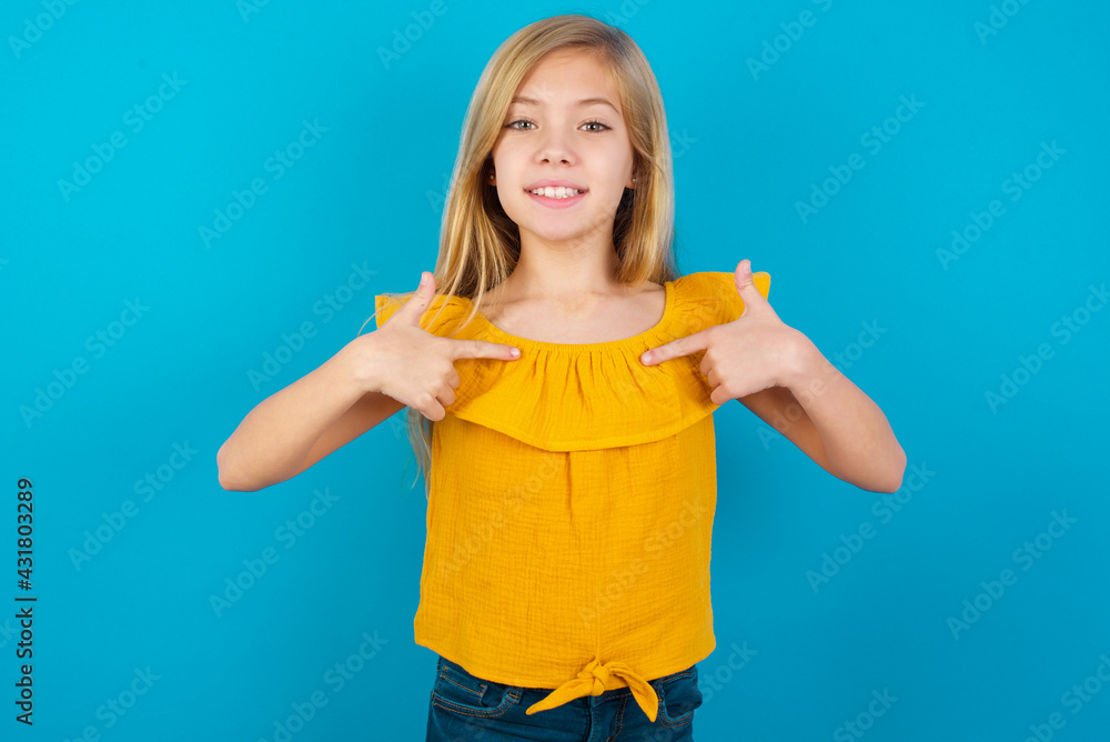 Caucasian kid girl wearing yellow T-shirt against blue wall points at his body, being in good mood after going shopping and making successful purchases Caucasian kid girl wearing yellow T-shirt against blue wall points at his body, being in good mood after going shopping and making successful purchases