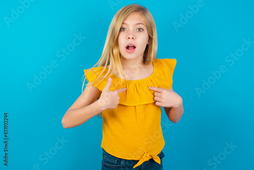 Embarrassed Caucasian kid girl wearing yellow T-shirt against blue indicates at herself with puzzled expression, being shocked to be chosen to participate in competition, hesitates about something