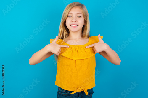 Caucasian kid girl wearing yellow T-shirt against blue wall points at his body, being in good mood after going shopping and making successful purchases