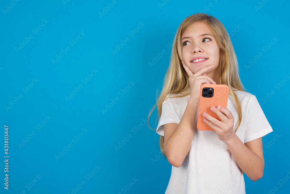 Image of a thinking dreaming beautiful Caucasian little girl wearing white T-shirt over blue background using mobile phone and holding hand on face. Taking decisions and social media concept. Image of a thinking dreaming beautiful Caucasian little girl wearing white T-shirt over blue background using mobile phone and holding hand on face. Taking decisions and social media concept.