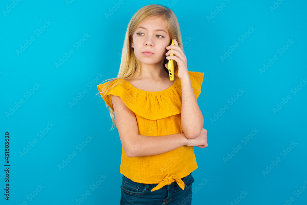Sad Caucasian kid girl wearing yellow T-shirt against blue wall talking on smartphone. Communication concept. Sad Caucasian kid girl wearing yellow T-shirt against blue wall talking on smartphone. Communication concept.