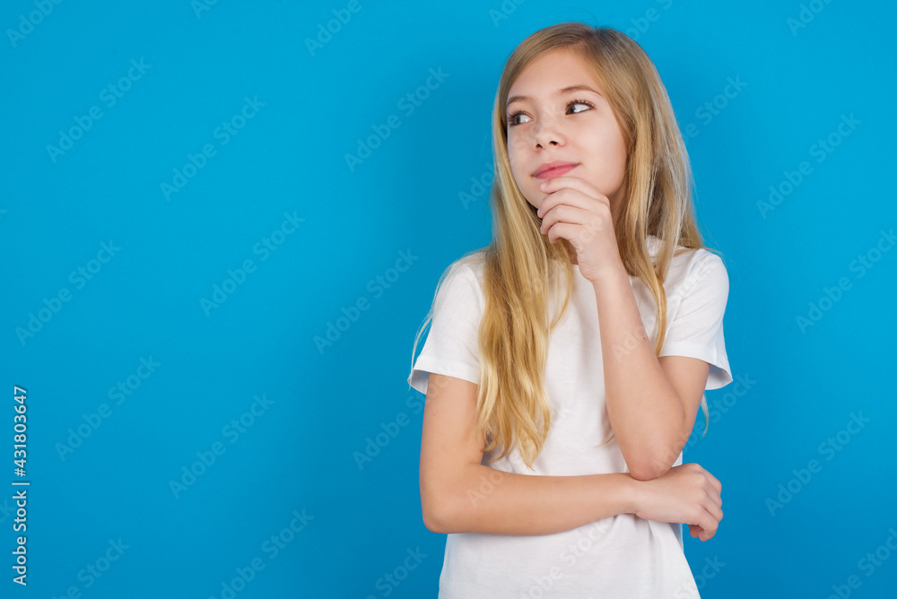 Thoughtful beautiful Caucasian little girl wearing white T-shirt over blue background holds chin and looks away pensively makes up great plan Thoughtful beautiful Caucasian little girl wearing white T-shirt over blue background holds chin and looks away pensively makes up great plan