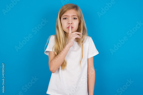 beautiful Caucasian little girl wearing white T-shirt over blue background makes silence gesture, keeps index finger to lips makes hush sign. Asks not to share secret.