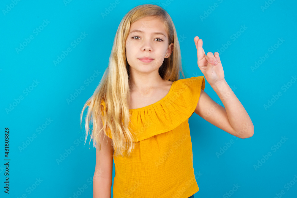 Fototapeta premium Caucasian kid girl wearing yellow T-shirt against blue wall pointing up with fingers number ten in Chinese sign language Shi