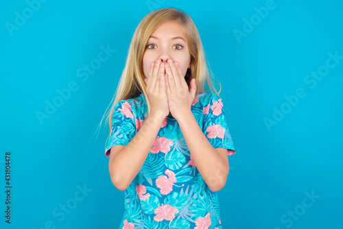 Caucasian kid girl wearing hawaiian T-shirt against blue wall keeps hands on mouth, looks with eyes full of disbelief, being puzzled with amount of work