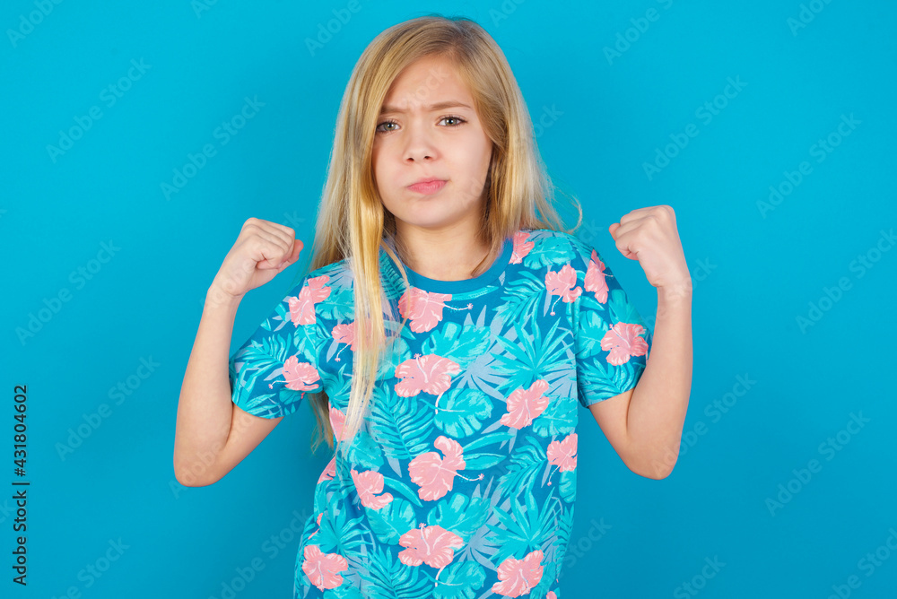 Irritated Caucasian kid girl wearing hawaiian T-shirt against blue wall blows cheeks with anger and raises clenched fists expresses rage and aggressive emotions. Furious model Irritated Caucasian kid girl wearing hawaiian T-shirt against blue wall blows cheeks with anger and raises clenched fists expresses rage and aggressive emotions. Furious model