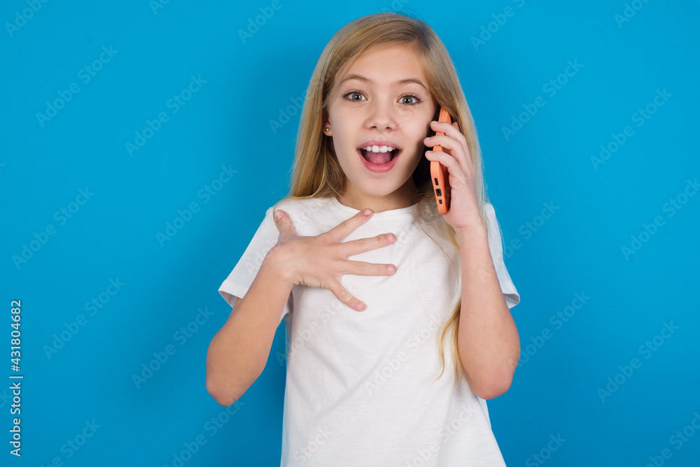 Smiling beautiful Caucasian little girl wearing white T-shirt over blue background talks via cellphone, enjoys pleasant great conversation. People, technology, communication concept Smiling beautiful Caucasian little girl wearing white T-shirt over blue background talks via cellphone, enjoys pleasant great conversation. People, technology, communication concept