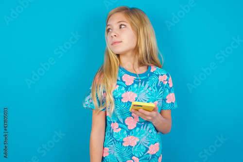 Caucasian kid girl wearing hawaiian T-shirt against blue wall holds mobile phone uses high speed internet and social networks has online communication. Modern technologies concept