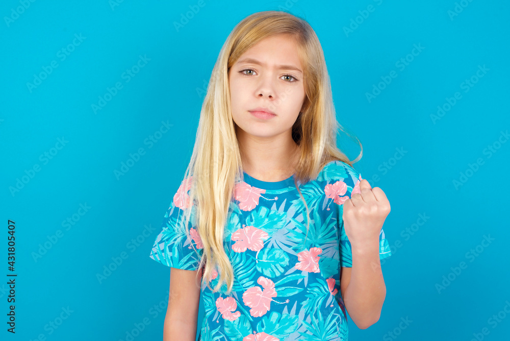 Caucasian kid girl wearing hawaiian T-shirt against blue wall shows fist has annoyed face expression going to revenge or threaten someone makes serious look. I will show you who is boss Caucasian kid girl wearing hawaiian T-shirt against blue wall shows fist has annoyed face expression going to revenge or threaten someone makes serious look. I will show you who is boss
