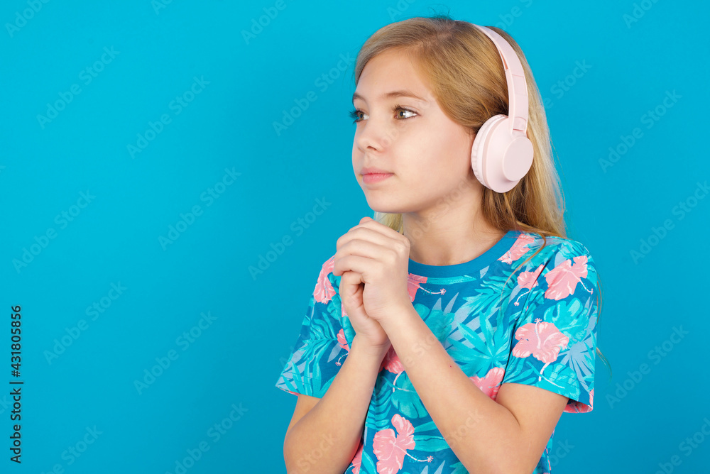 Caucasian kid girl wearing hawaiian T-shirt against blue wall wears stereo headphones listening to music concentrated and looking aside with interest. Caucasian kid girl wearing hawaiian T-shirt against blue wall wears stereo headphones listening to music concentrated and looking aside with interest.
