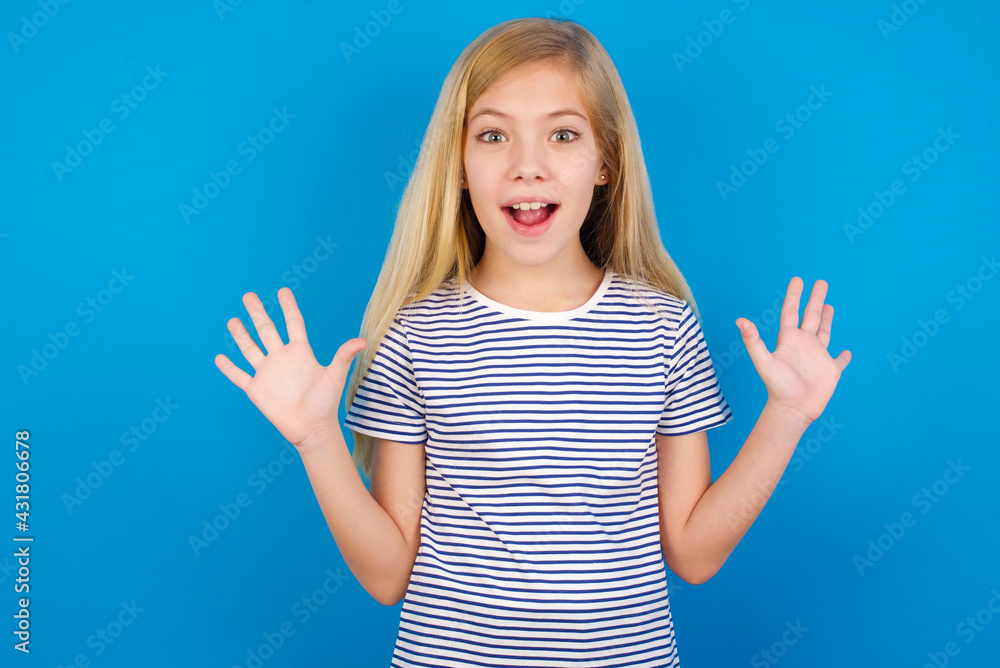 Fototapeta premium Optimistic Caucasian kid girl wearing striped shirt ​against blue wall raises palms from joy, happy to receive awesome present from someone, shouts loudly, Excited model screaming.