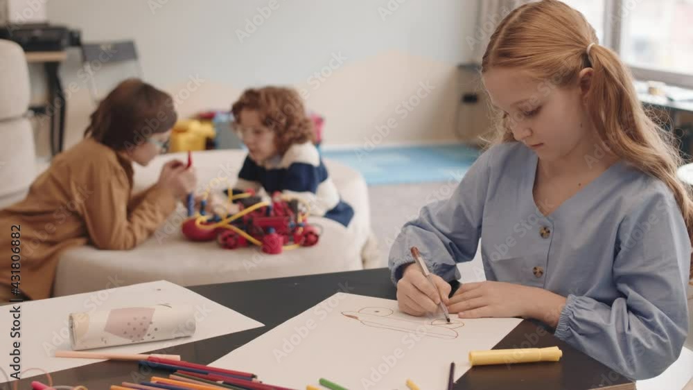 Lockdown of red-haired Caucasian schoolgirl sitting at desktop in classroom and drawing while her two male classmates constructing robot on background Lockdown of red-haired Caucasian schoolgirl sitting at desktop in classroom and drawing while her two male classmates constructing robot on background