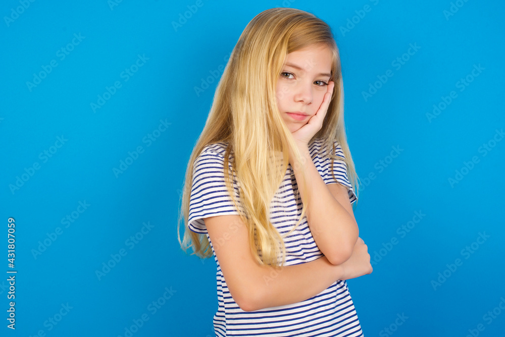 Very bored Caucasian kid girl wearing striped shirt against blue wall holding hand on cheek while support it with another crossed hand, looking tired and sick, Very bored Caucasian kid girl wearing striped shirt against blue wall holding hand on cheek while support it with another crossed hand, looking tired and sick,