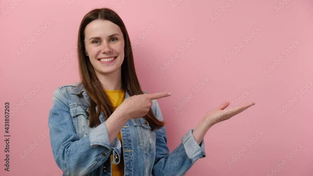 Cute smiling young woman pointing fingers hands aside on workspace copy space mockup promo commercial area isolated on pink studio background wall, wears denim jacket. People emotion lifestyle concept Cute smiling young woman pointing fingers hands aside on workspace copy space mockup promo commercial area isolated on pink studio background wall, wears denim jacket. People emotion lifestyle concept