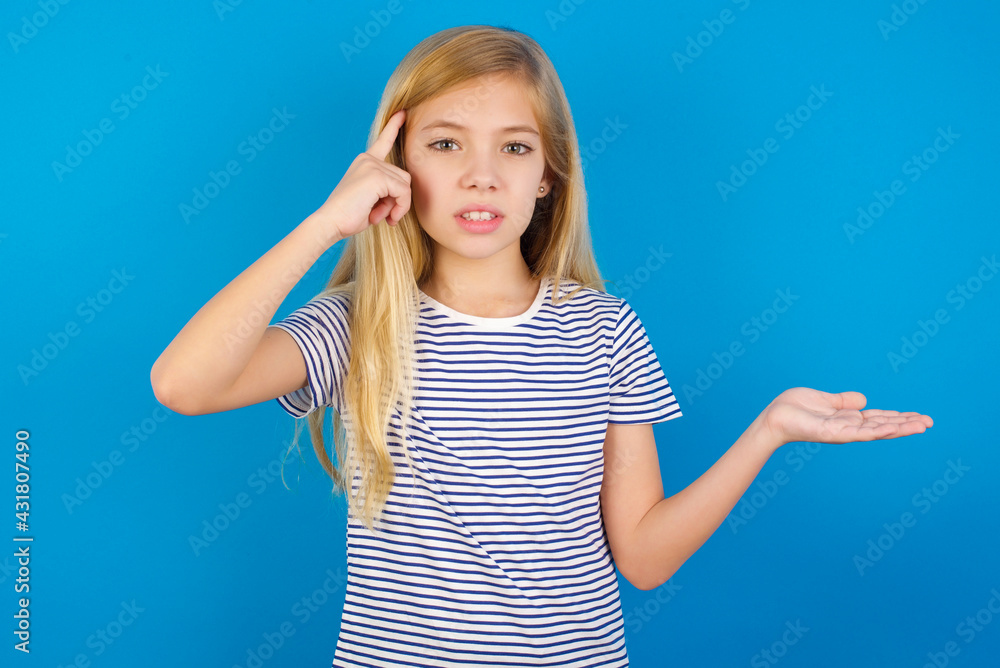 Fototapeta premium Caucasian kid girl wearing striped shirt ​against blue wall confused and annoyed with open palm showing copy space and pointing finger to forehead. Think about it.