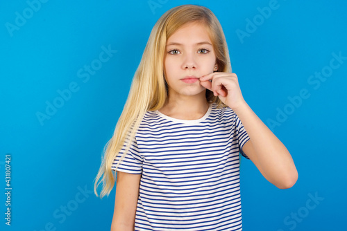Caucasian kid girl wearing striped shirt ​against blue wall  mouth and lips shut as zip with fingers. Secret and silent, taboo talking.