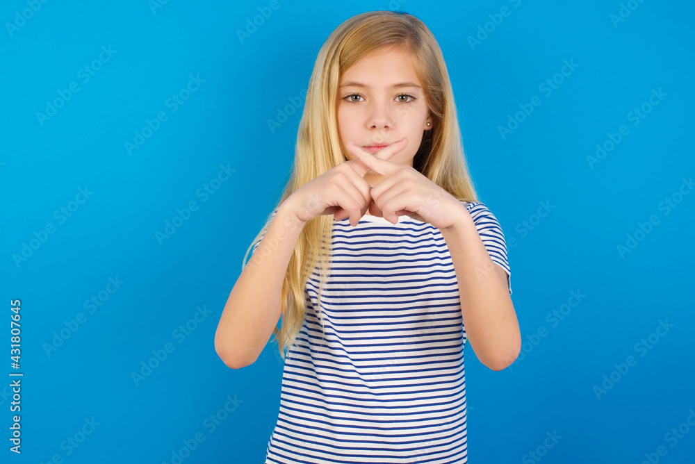 Caucasian kid girl wearing striped shirt against blue wall Has rejection angry expression crossing fingers doing negative sign. Caucasian kid girl wearing striped shirt against blue wall Has rejection angry expression crossing fingers doing negative sign.