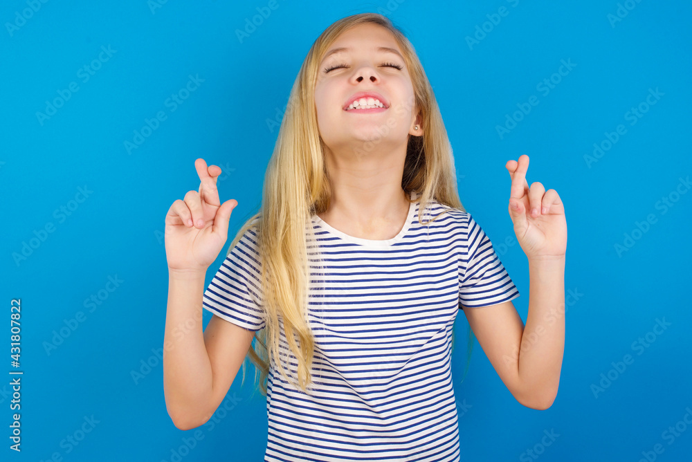 Caucasian kid girl wearing striped shirt against blue wall gesturing finger crossed smiling with hope and eyes closed. Luck and superstitious concept. Caucasian kid girl wearing striped shirt against blue wall gesturing finger crossed smiling with hope and eyes closed. Luck and superstitious concept.