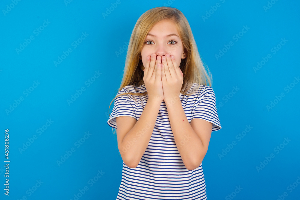 Vivacious Caucasian kid girl wearing striped shirt against blue wall , giggles joyfully, covers mouth, has natural laughter, hears positive story or funny anecdote Vivacious Caucasian kid girl wearing striped shirt against blue wall , giggles joyfully, covers mouth, has natural laughter, hears positive story or funny anecdote