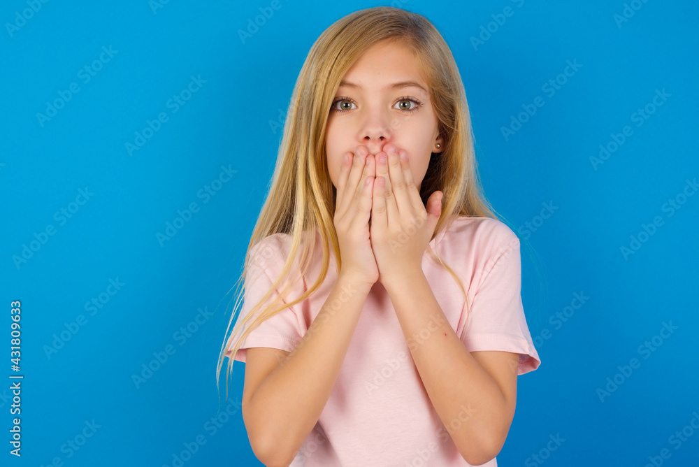 Caucasian kid girl wearing pink shirt against blue wall keeps hands on mouth, looks with eyes full of disbelief, being puzzled with amount of work Caucasian kid girl wearing pink shirt against blue wall keeps hands on mouth, looks with eyes full of disbelief, being puzzled with amount of work