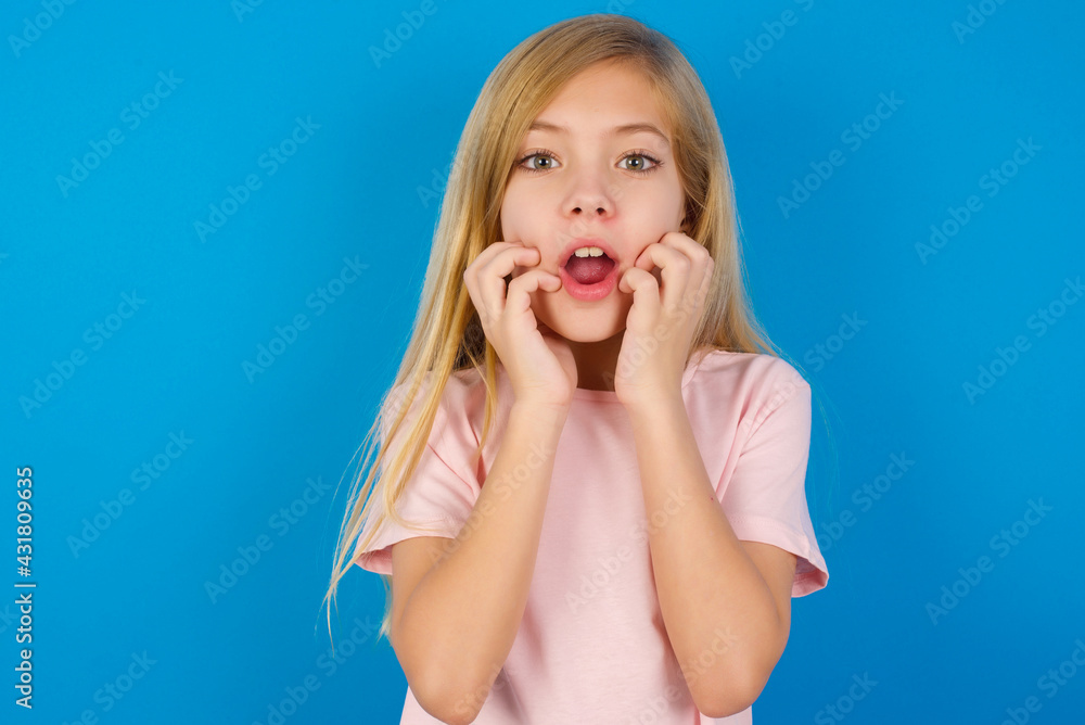 Speechless Caucasian kid girl wearing pink shirt against blue wall keeps hands near opened mouth reacts to shocking news stares wondered at camera Speechless Caucasian kid girl wearing pink shirt against blue wall keeps hands near opened mouth reacts to shocking news stares wondered at camera