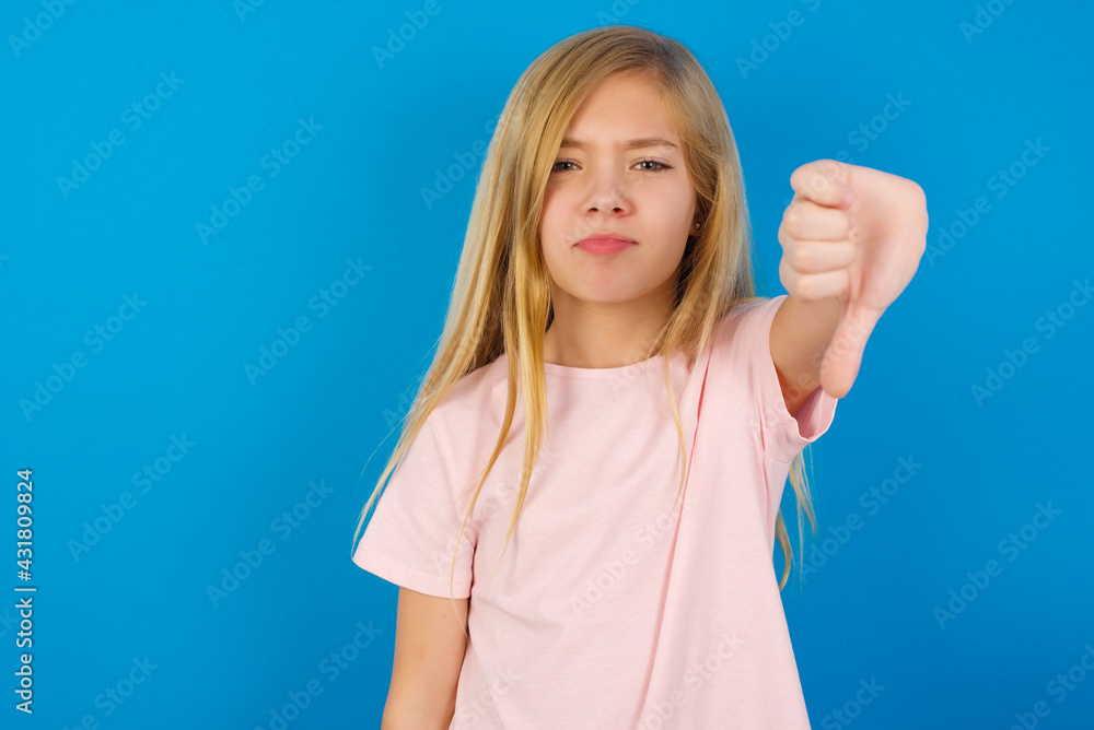 Fototapeta premium Caucasian kid girl wearing pink shirt against blue wall looking unhappy and angry showing rejection and negative with thumbs down gesture. Bad expression.