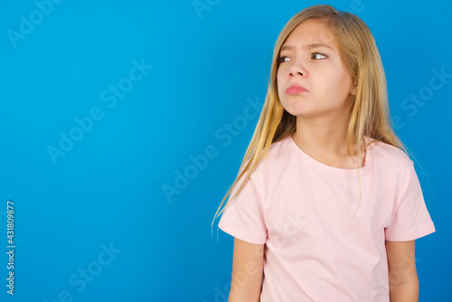 Dissatisfied Caucasian kid girl wearing pink shirt against blue wall purses lips and has unhappy expression looks away stands offended. Depressed frustrated model. © Roquillo