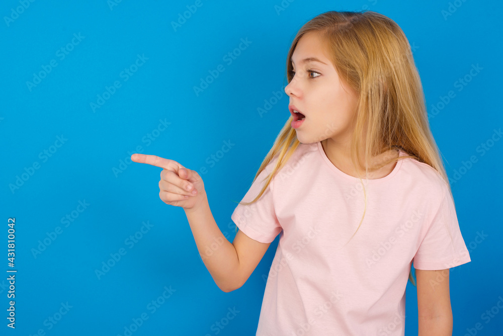 Stunned Caucasian kid girl wearing pink shirt against blue wall with greatly surprised expression points away on copy space, indicates something Stunned Caucasian kid girl wearing pink shirt against blue wall with greatly surprised expression points away on copy space, indicates something