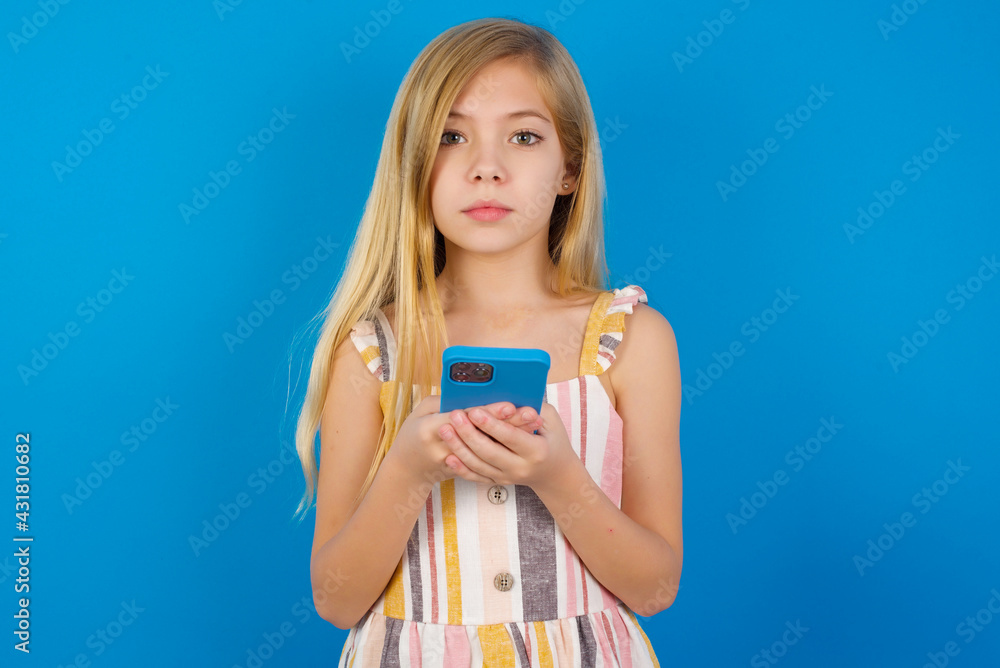 Portrait of serious confident Caucasian kid girl wearing dress against blue wall holding phone in two hands Portrait of serious confident Caucasian kid girl wearing dress against blue wall holding phone in two hands