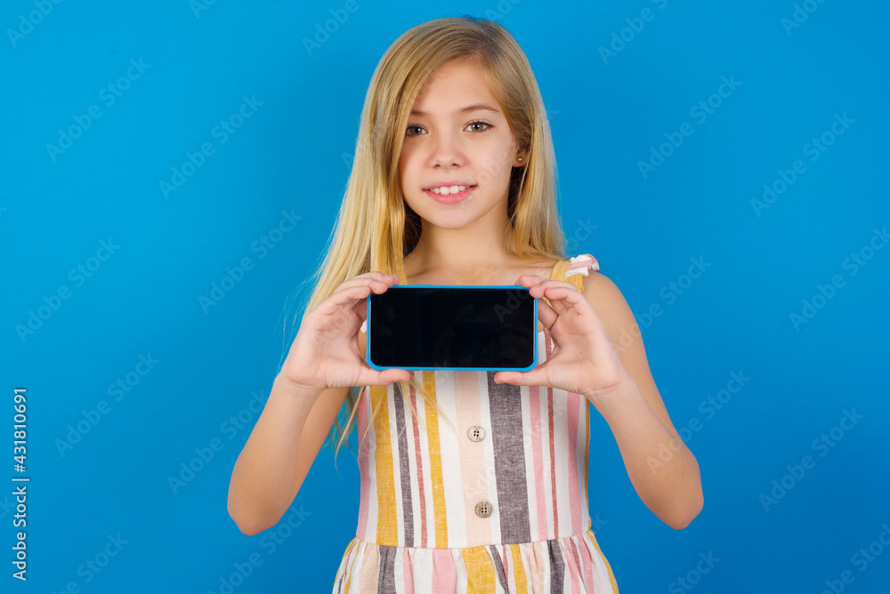 Cheerful cheery content Caucasian kid girl wearing dress against blue wall holding in hands device hobby smm post blog Cheerful cheery content Caucasian kid girl wearing dress against blue wall holding in hands device hobby smm post blog