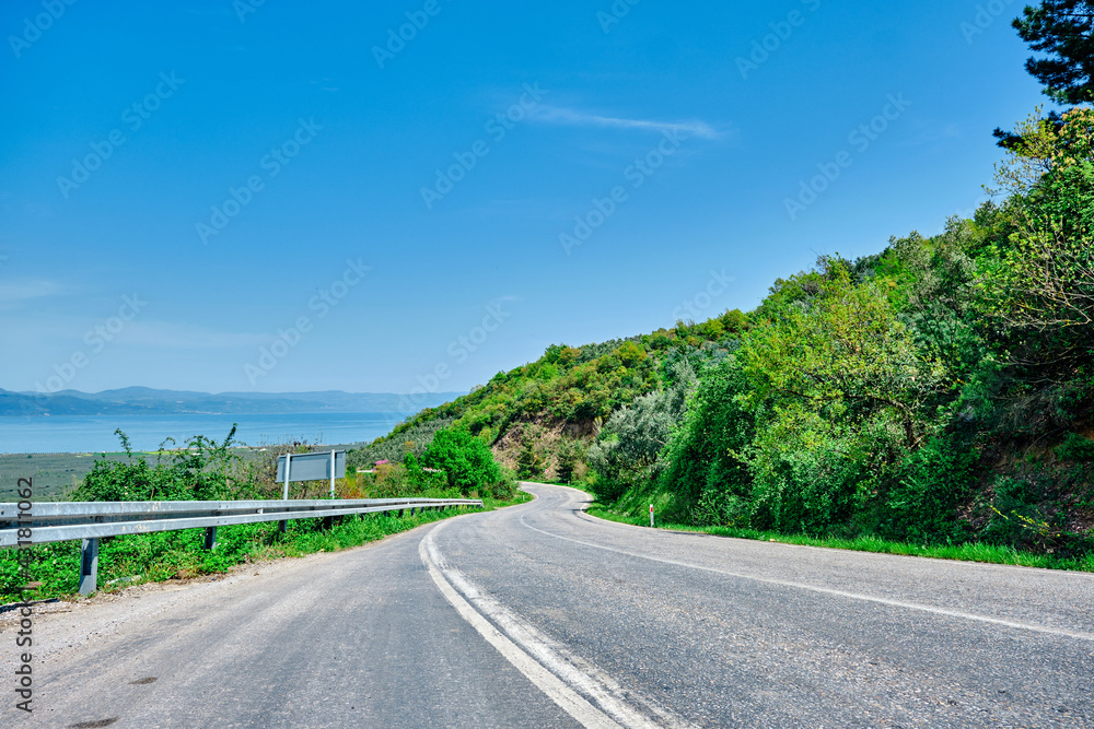 an asphalt road to Bursa to Nicaea (iznik) during sunny day and road signs and protection fences next to the road with small hills that is covered by many green plants and grass with lake of Nicaea.