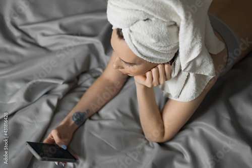 Pretty young woman with a towel on her head lies on bed hold mobile phone and check the social networks. Remote work, online shopping, quarantine isolation