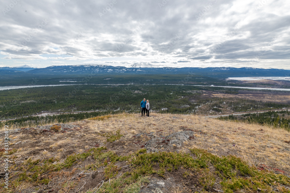 Two people, couple hikers standing on a panoramic view above Yukon River outside of Whitehorse in north of Canada. Boreal forest, snow capped mountains on a cloudy afternoon in distance. Two people, couple hikers standing on a panoramic view above Yukon River outside of Whitehorse in north of Canada. Boreal forest, snow capped mountains on a cloudy afternoon in distance.
