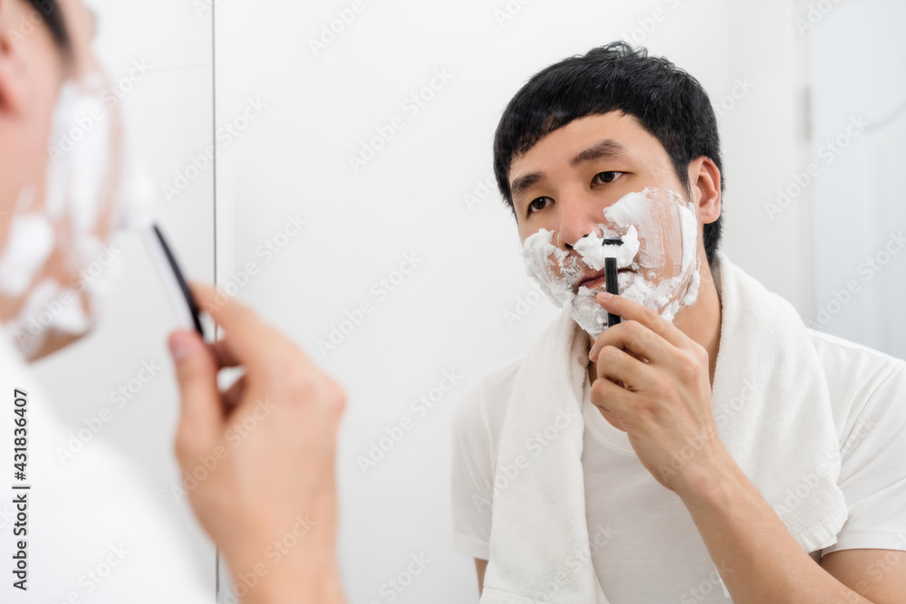 man using razor to shaving his face with cream foam in bathroom mirror