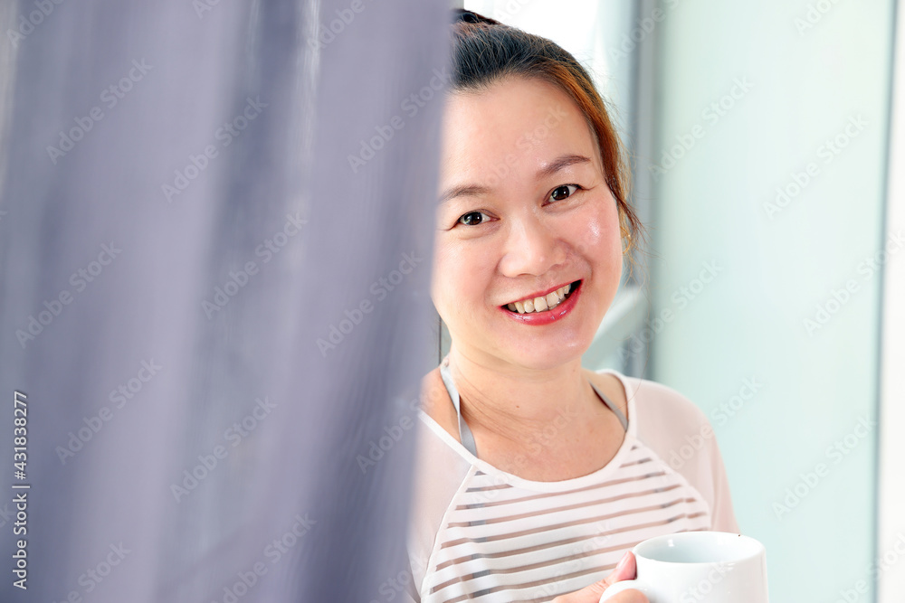 Beautiful attractive middle age southeast Asian woman standing next to daylight window curtain drink coffee from cup look at camera Beautiful attractive middle age southeast Asian woman standing next to daylight window curtain drink coffee from cup look at camera