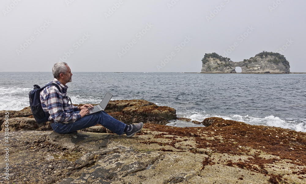 gray-haired man in jeans and a plaid shirt works with a laptop on the ocean shore